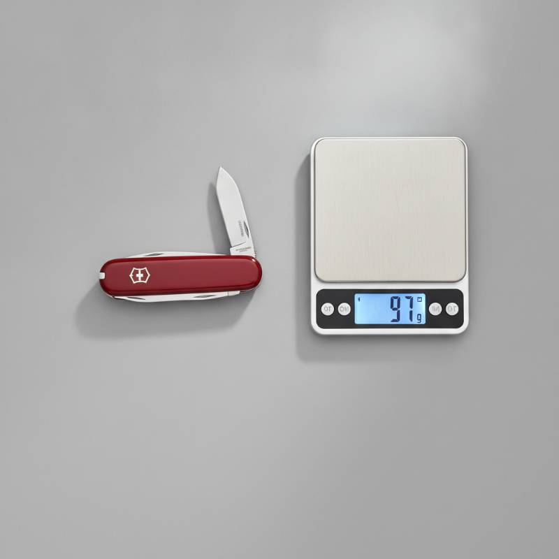 A top-down view of a closed red Victorinox Huntsman next to a tape measure and a digital pocket scale showing its weight