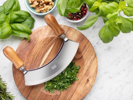A traditional mezzaluna knife and ulu knife on a wooden cutting board with fresh herbs